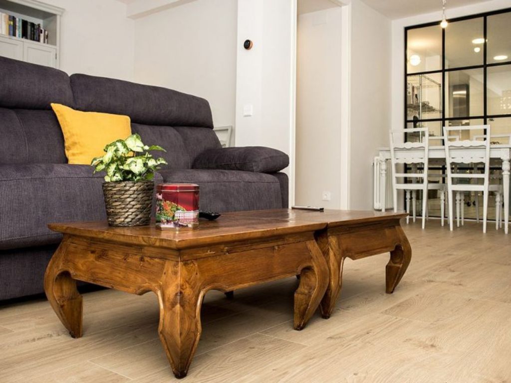 Mesa de madera con decoración, sofá gris y comedor al fondo.